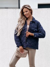Woman wearing a navy blue quilted jacket with beige pants and a clutch against a light background