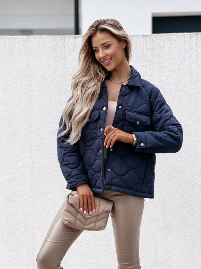 Woman wearing a navy blue quilted jacket with beige pants and a clutch against a light background