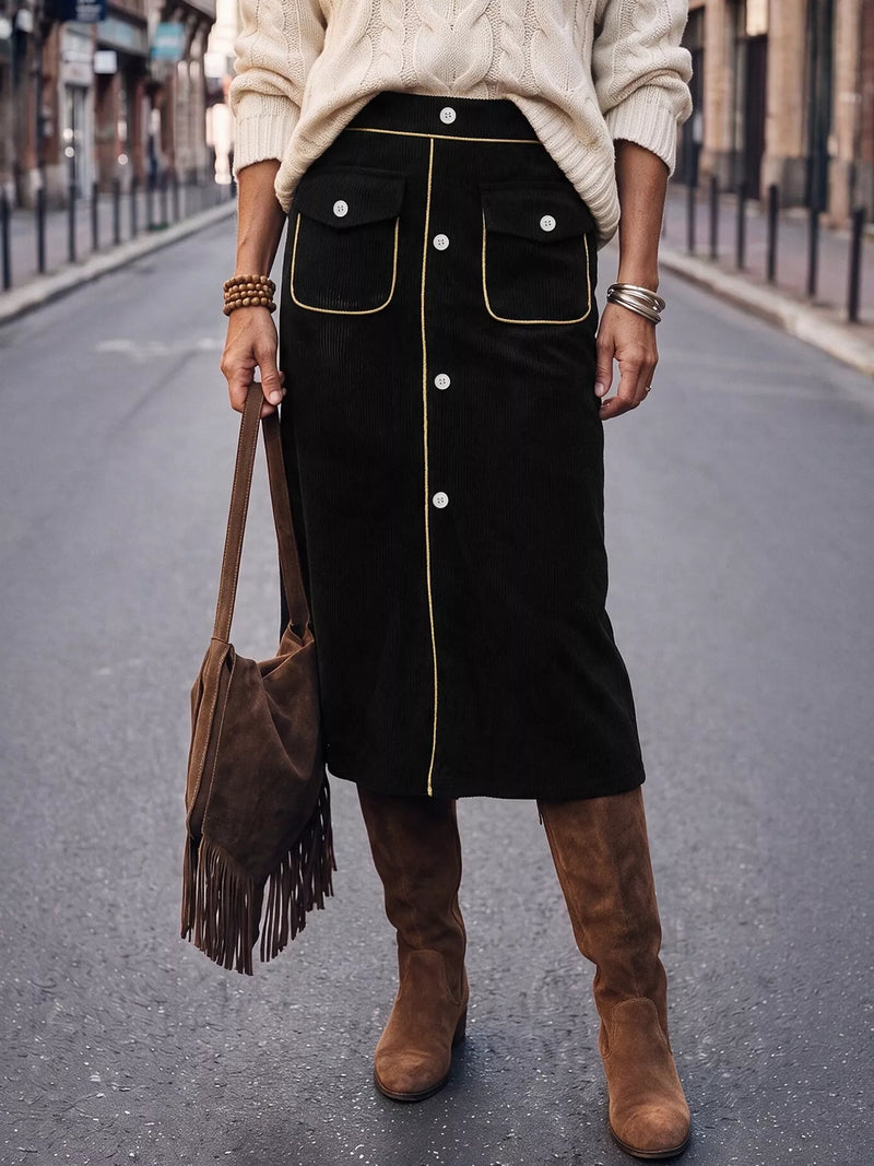 A person standing on a city street wearing a black midi skirt with gold trim and white buttons, a cream cable-knit sweater, brown suede knee-high boots, and carrying a brown fringe shoulder bag.
