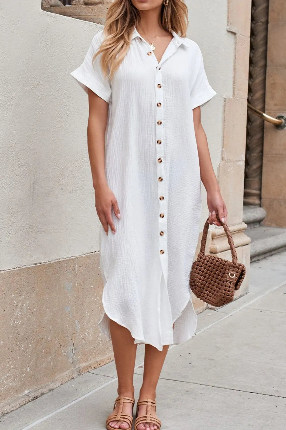 Woman wearing a white button-up dress with a brown handbag against a beige wall.