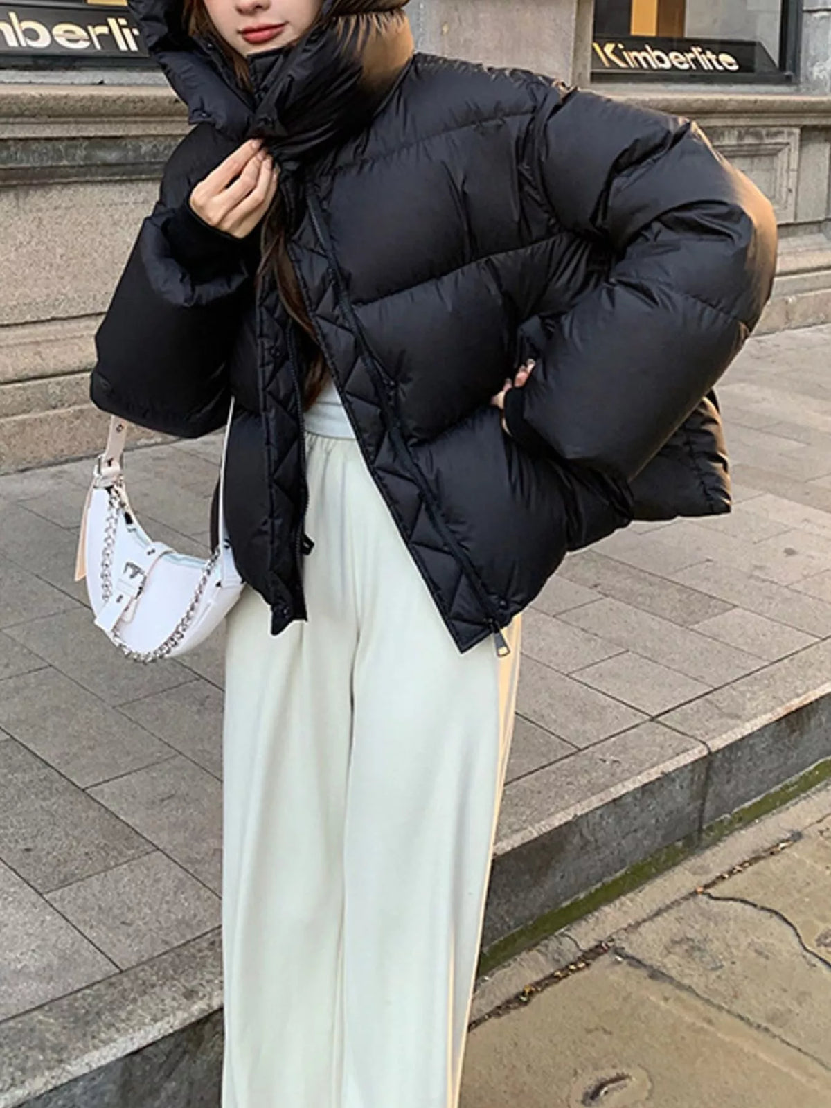 A person wearing a black puffer jacket with a high collar, paired with cream wide-leg pants and a white shoulder bag, standing outdoors on stone steps.