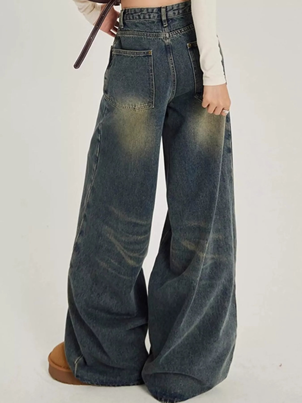 A person wearing very wide-leg faded blue denim jeans with a dark wash and yellowed faded areas, paired with a white long-sleeve top and brown shoes, shown from the waist down against a plain light background.