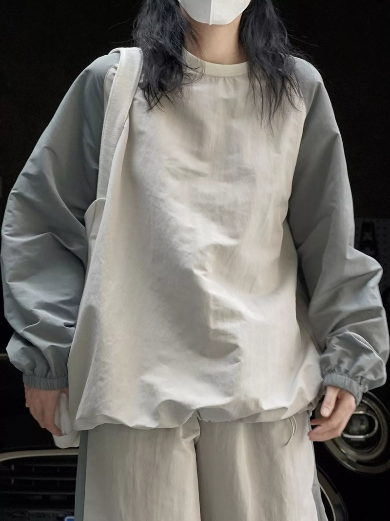 A person wearing an oversized light beige linen-blend top with contrasting light gray sleeves, paired with matching beige pants and a white shoulder bag, set against a dark background.