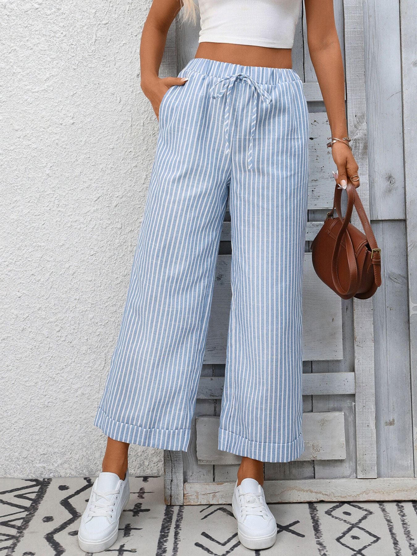 Person wearing blue and white striped wide-leg pants with a white top and white sneakers, standing against a light gray wall.