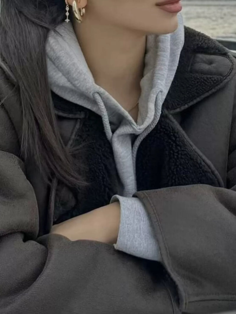 A close-up lifestyle shot of a person wearing a dark brown shearling-lined coat over a light gray hoodie, with dark hair and gold hoop earrings visible.