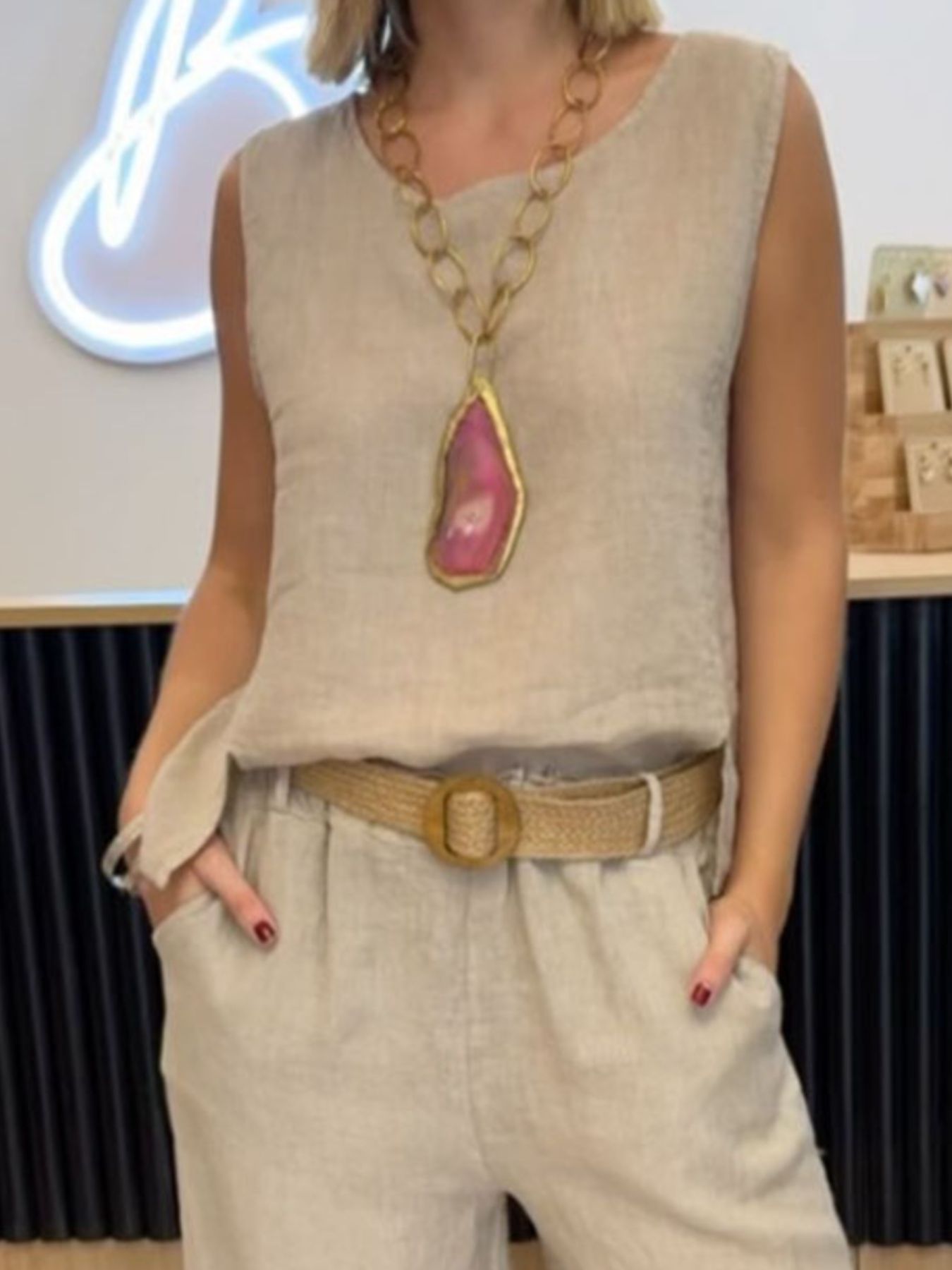 Person wearing a beige sleeveless top with a large necklace and beige pants in an indoor setting.