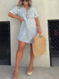 Woman wearing a light blue denim dress holding a straw bag against a plain wall.