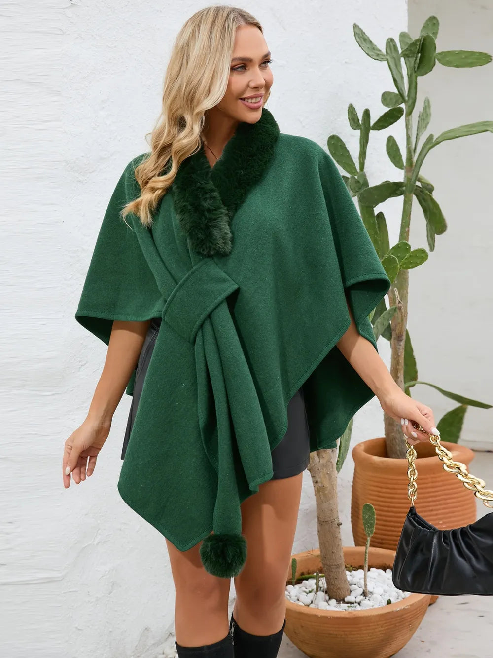 Woman wearing a green poncho with fur trim standing next to a potted plant.