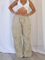 A model wearing beige wide-leg cargo pants with a drawstring waist and side pockets, paired with a white crop top, standing in front of a white draped fabric backdrop.