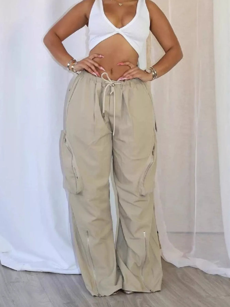 A model wearing beige wide-leg cargo pants with a drawstring waist and side pockets, paired with a white crop top, standing in front of a white draped fabric backdrop.