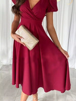 Woman wearing a burgundy dress holding a clutch against a white curtain background