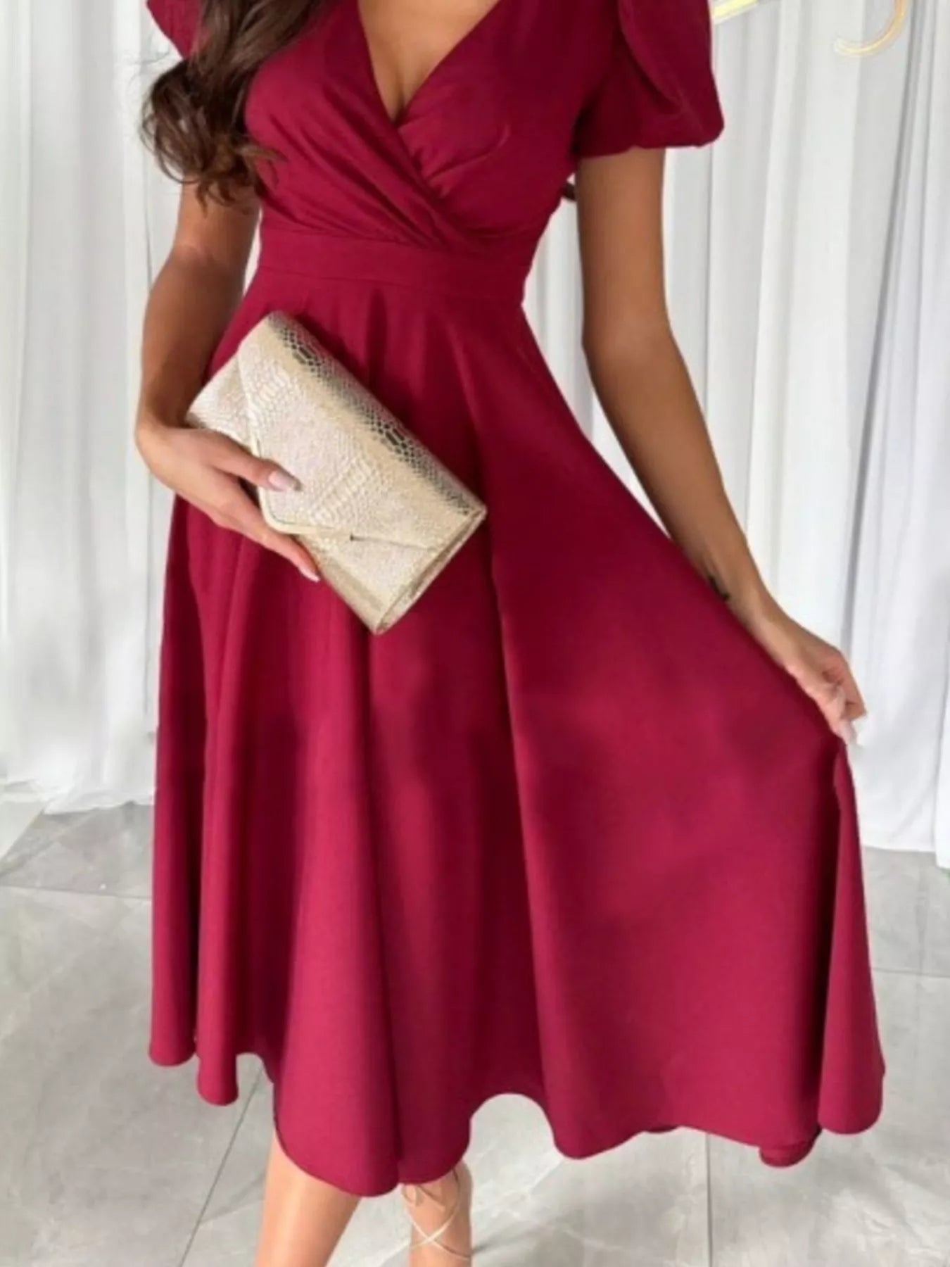 Woman wearing a burgundy dress holding a clutch against a white curtain background
