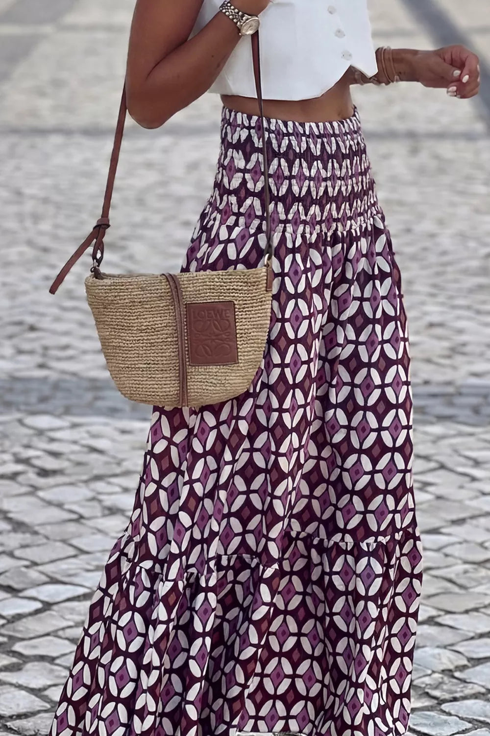 Person wearing a patterned skirt and holding a straw bag on a cobblestone street.
