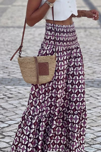 Person wearing a patterned skirt and holding a straw bag on a cobblestone street.