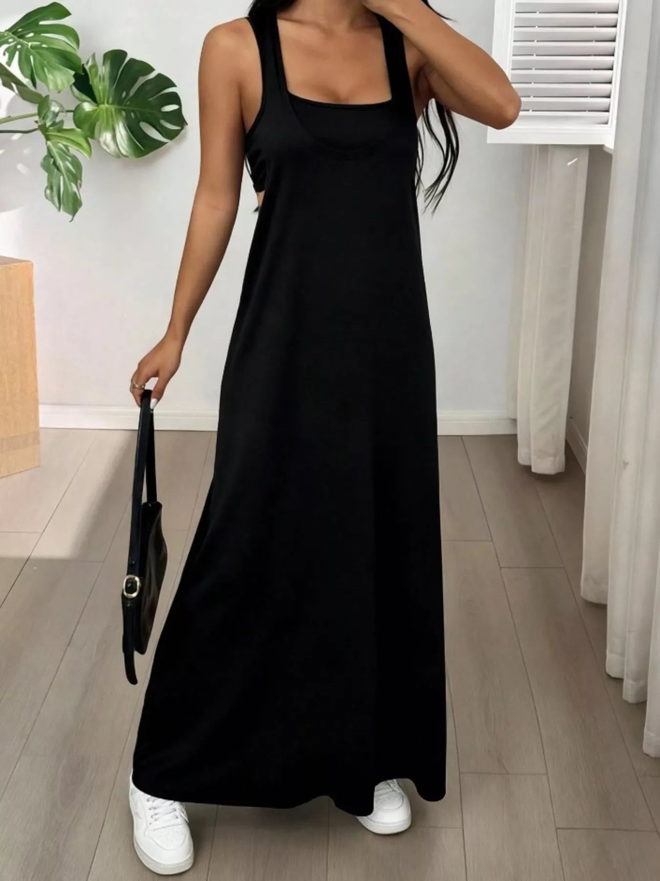 Woman wearing a black sleeveless dress holding a black handbag indoors.