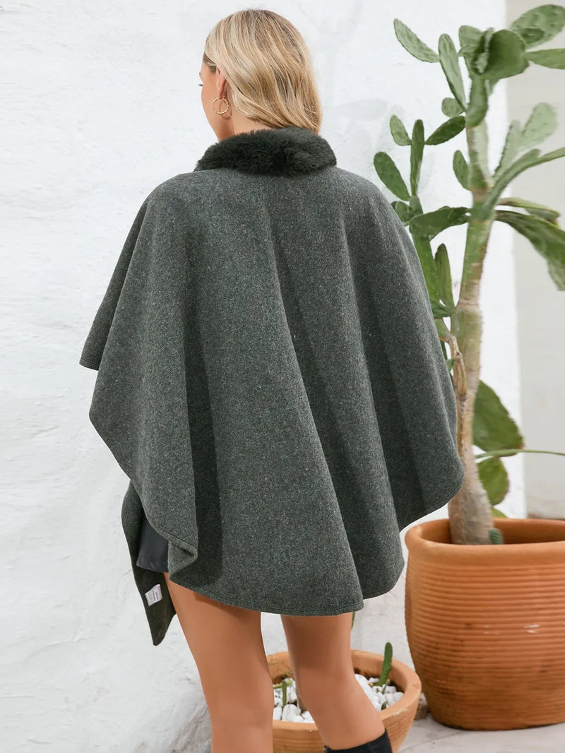 Person wearing a dark gray poncho with a fur collar, standing indoors next to a potted plant.