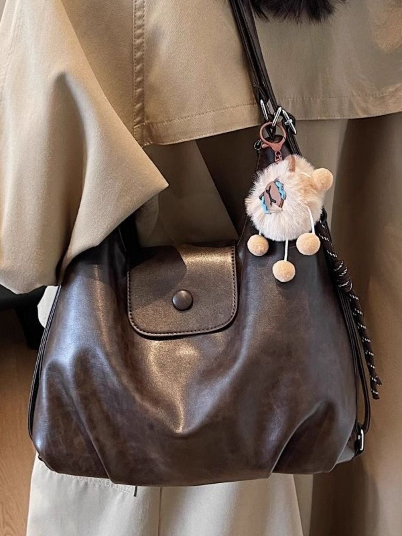 Brown leather handbag with a fluffy pom-pom charm on a person wearing a beige jacket.
