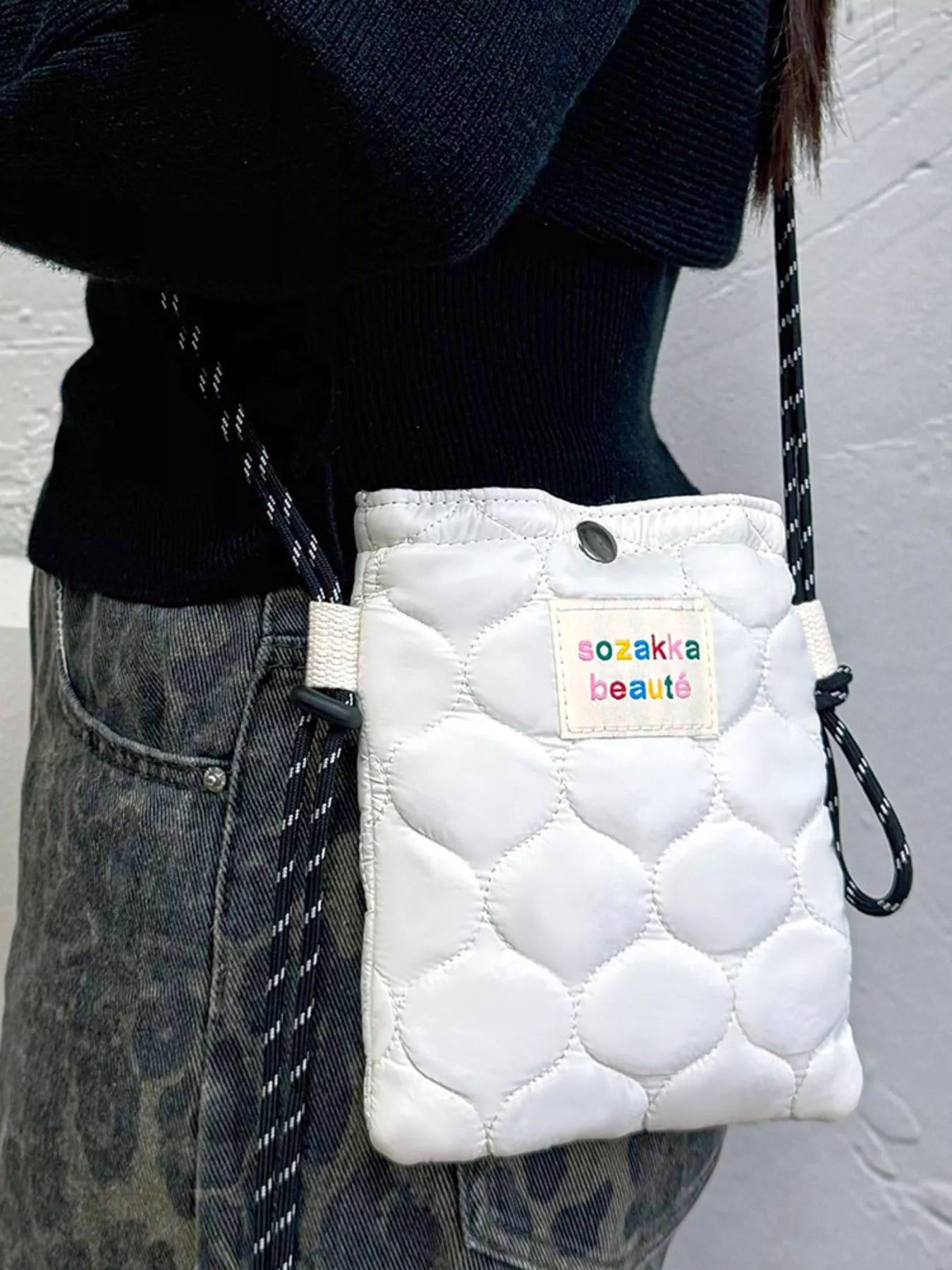 White quilted pouch with 'sozakia beauty' label held by a person wearing a black sweater and denim jeans.