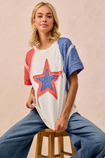 Woman wearing a white t-shirt with a red and blue star design, sitting on a wooden stool against a beige background.
