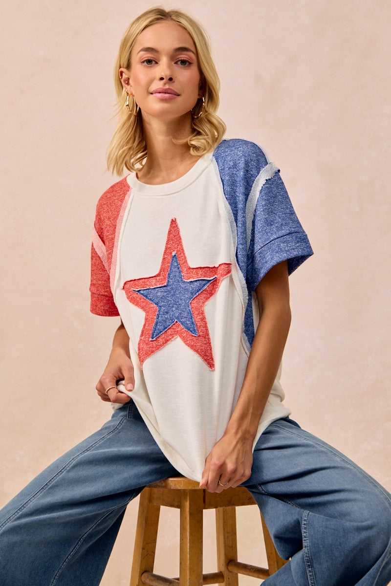 Woman wearing a white t-shirt with a red and blue star design, sitting on a wooden stool against a beige background.