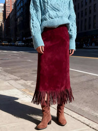 Person wearing a blue cable knit sweater and red suede skirt with fringe on a city street.