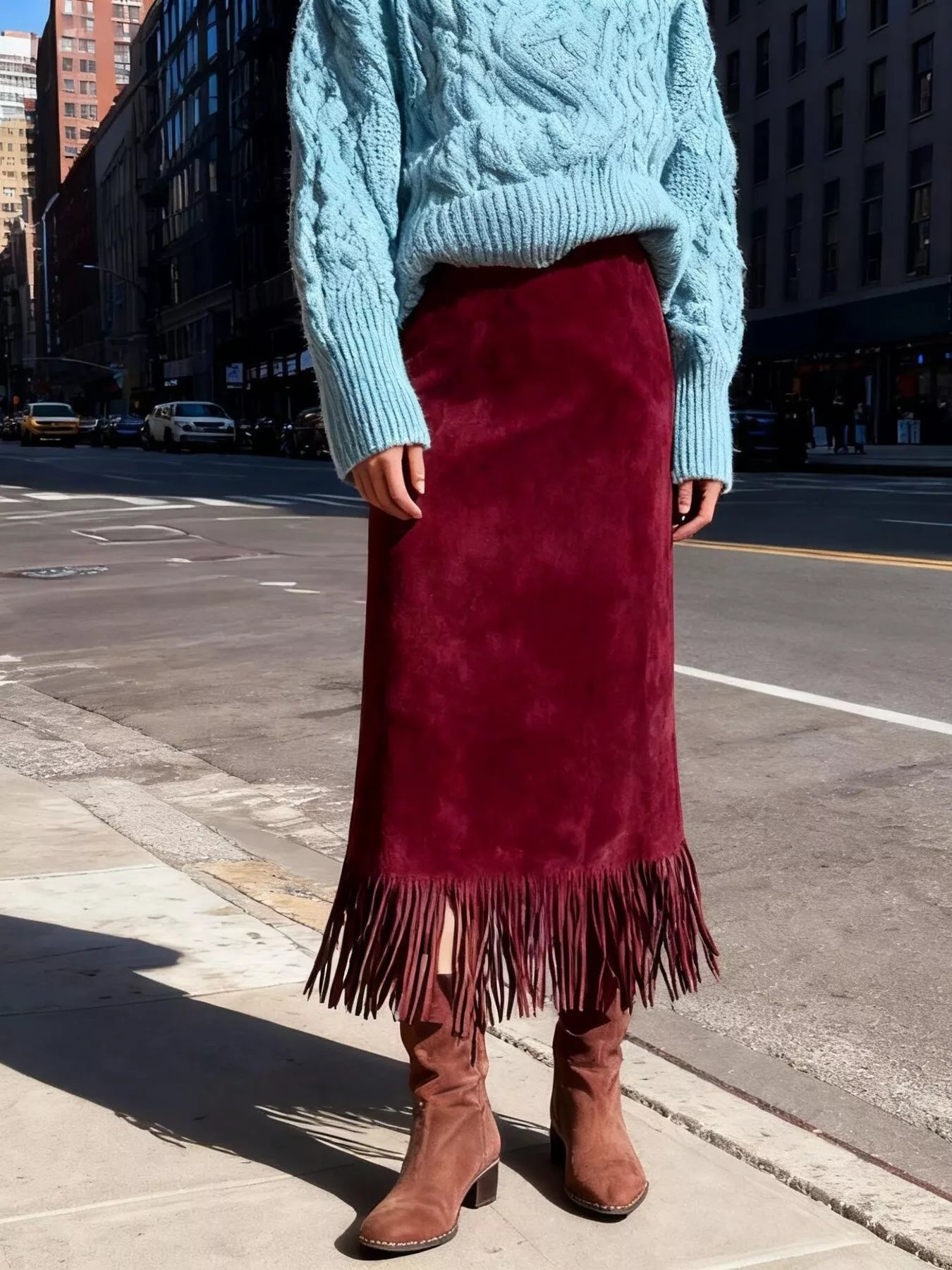 Person wearing a blue cable knit sweater and red suede skirt with fringe on a city street.