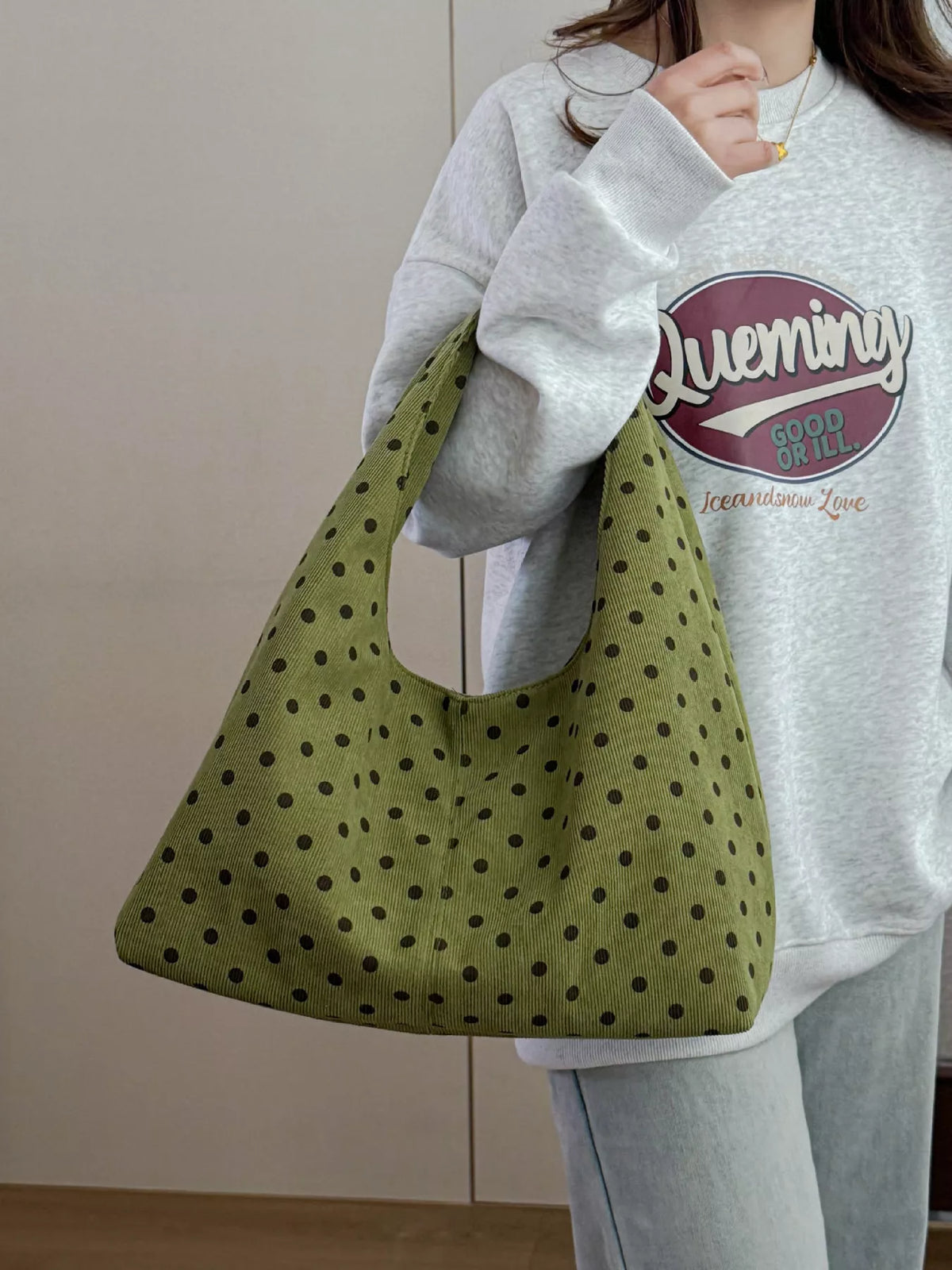A person wearing a light gray sweatshirt and jeans carries a large olive green fabric tote bag with black polka dots. The sweatshirt features a circular graphic with the text 'Queming' and 'Good to All'.