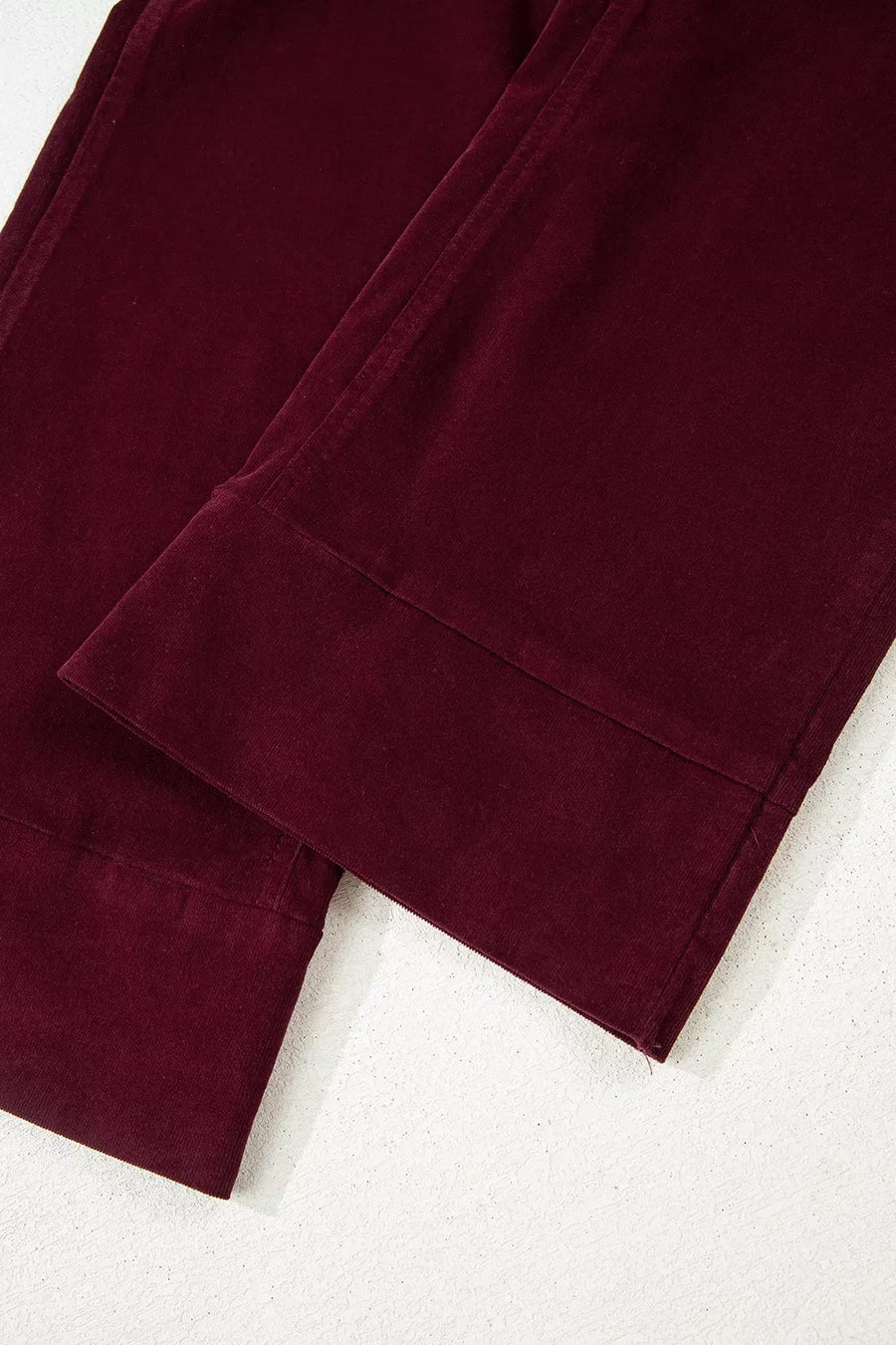 A close-up of dark burgundy corduroy pants folded on a light gray textured surface, showing the ribbed fabric texture and cuff construction.