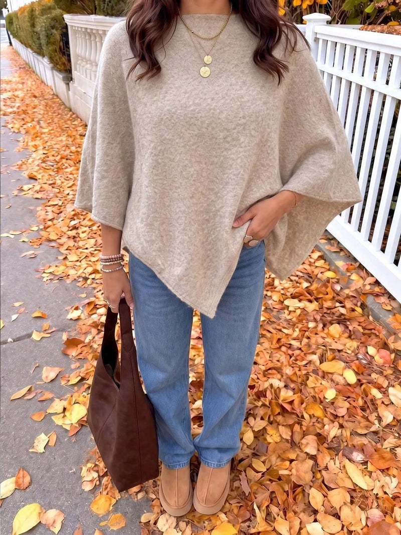 A woman wearing a beige knit poncho sweater, blue jeans, and tan boots stands on a sidewalk covered with fallen autumn leaves, holding a brown leather tote bag.