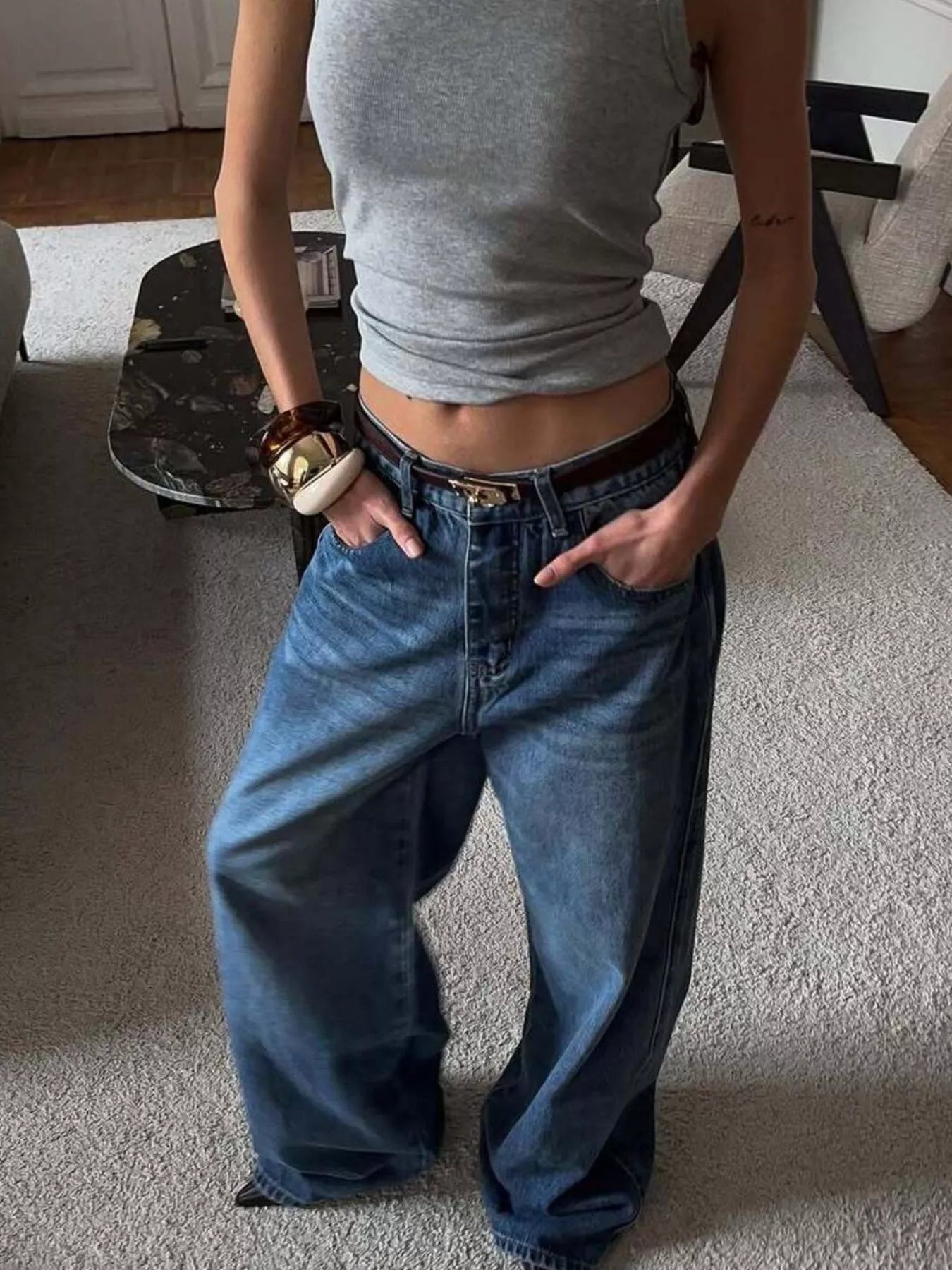 Person wearing a gray tank top and blue jeans standing on a carpeted floor.