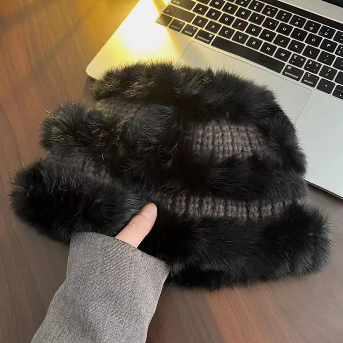 Black furry slippers held by a hand in front of a laptop on a wooden surface