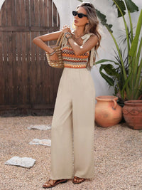Woman in a colorful top and beige pants standing outdoors with plants and a wooden structure in the background.