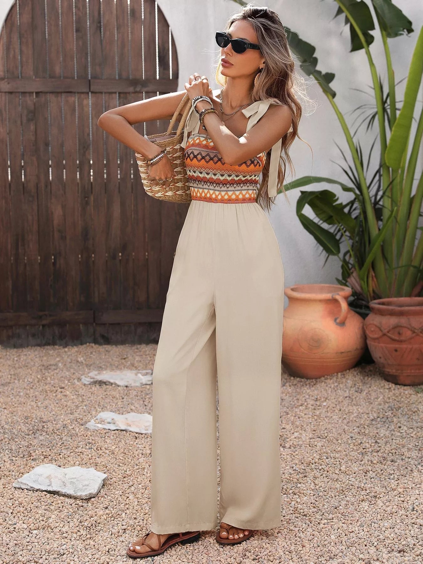 Woman in a colorful top and beige pants standing outdoors with plants and a wooden structure in the background.