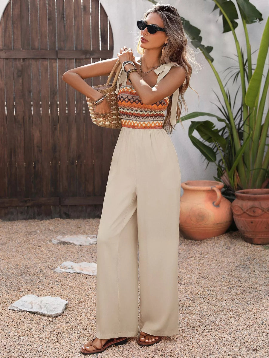 Woman in a colorful top and beige pants standing outdoors with plants and a wooden structure in the background.