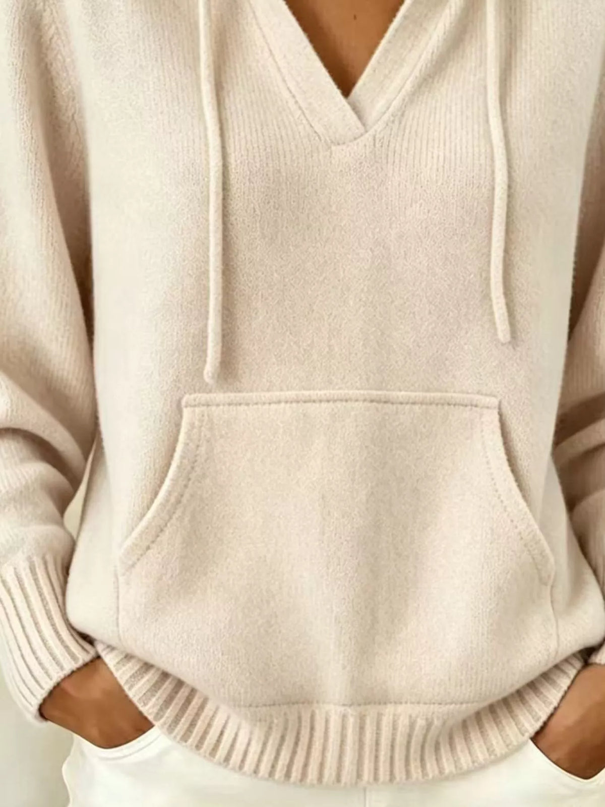 A close-up of a person wearing a soft knit pullover hoodie in a light cream or oatmeal color. The hoodie features a V-neck, long sleeves with ribbed cuffs, a large front kangaroo pocket, and ribbed hem.