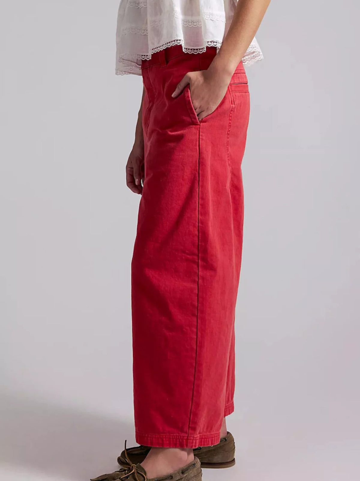 A person wearing a bright red wide-leg skirt or pants with side pockets, paired with a white lace-trim top and brown shoes, shown against a light gray studio background.