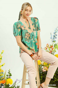 Woman wearing a floral blouse sitting on a stool with flowers around her.