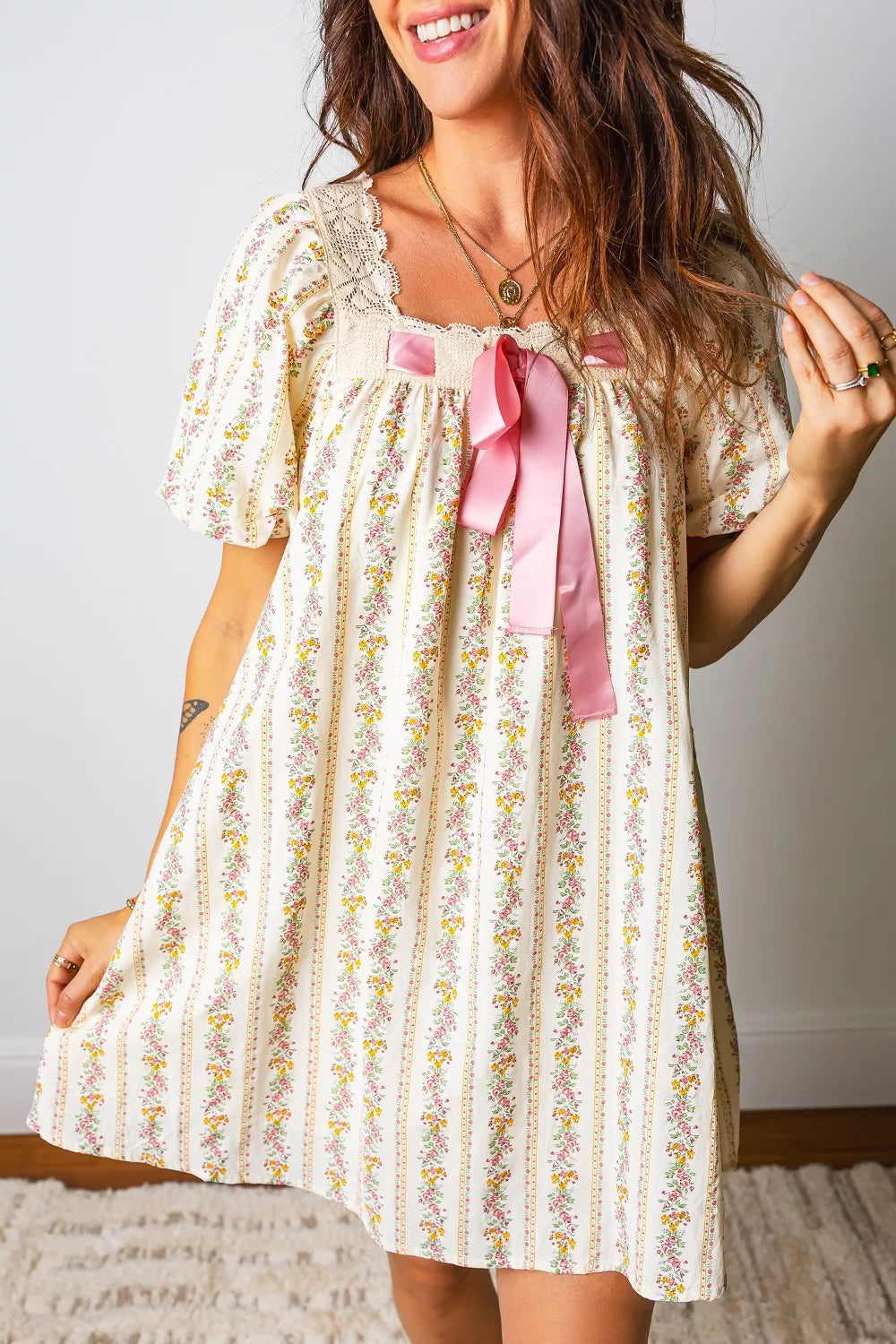 Woman wearing a floral dress with a pink ribbon bow, standing against a plain background.