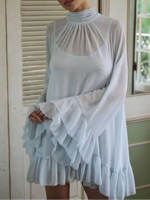 A woman wearing a light blue high-neck long-sleeve blouse with dramatic layered ruffle sleeves and a ruffled hem, shown in a softly lit indoor setting.