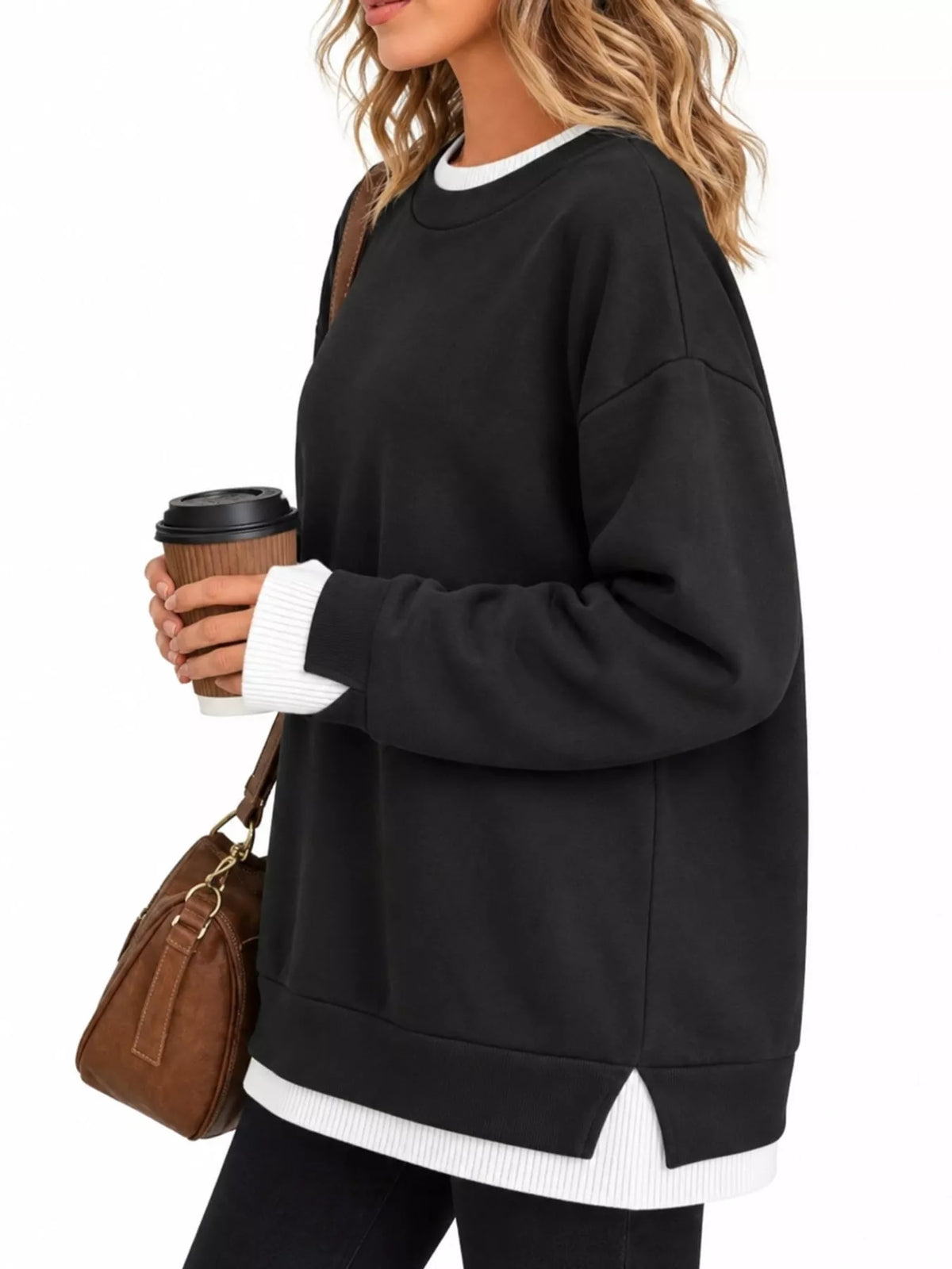 A woman wearing an oversized black crewneck sweatshirt with white ribbed trim at the collar, cuffs, and hem, holding a brown paper coffee cup and wearing a brown shoulder bag against a plain white background.