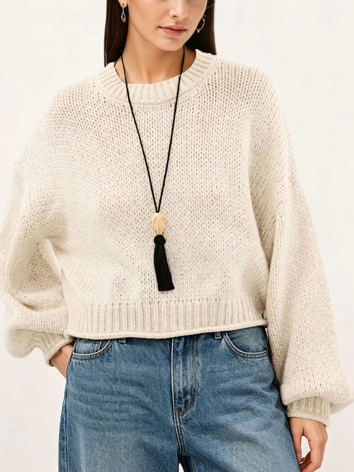 A woman wearing a cream-colored, textured knit sweater with balloon sleeves and blue jeans, accessorized with a long black tassel necklace.