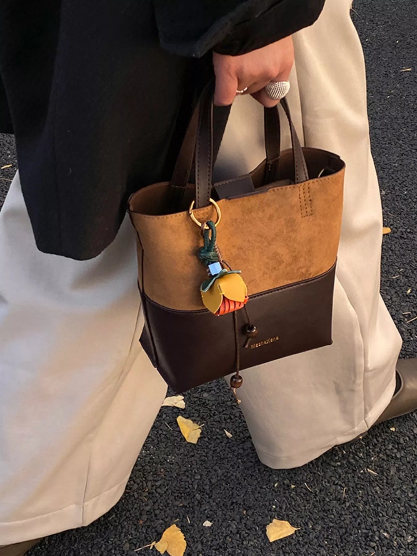 Person holding a brown and black handbag with a decorative charm on an outdoor surface.