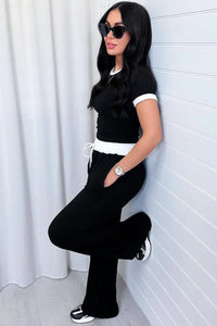 Woman in black outfit with white accents leaning against a white wall.