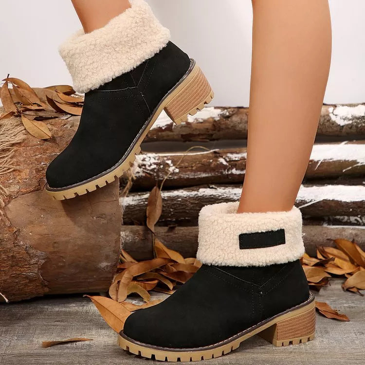 Black ankle boots with white fur trim worn by a person, standing on a wooden log with autumn leaves.