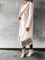 Person wearing a long beige coat and white boots, holding a matching handbag against a gray concrete wall.