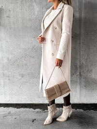 Person wearing a long beige coat and white boots, holding a matching handbag against a gray concrete wall.