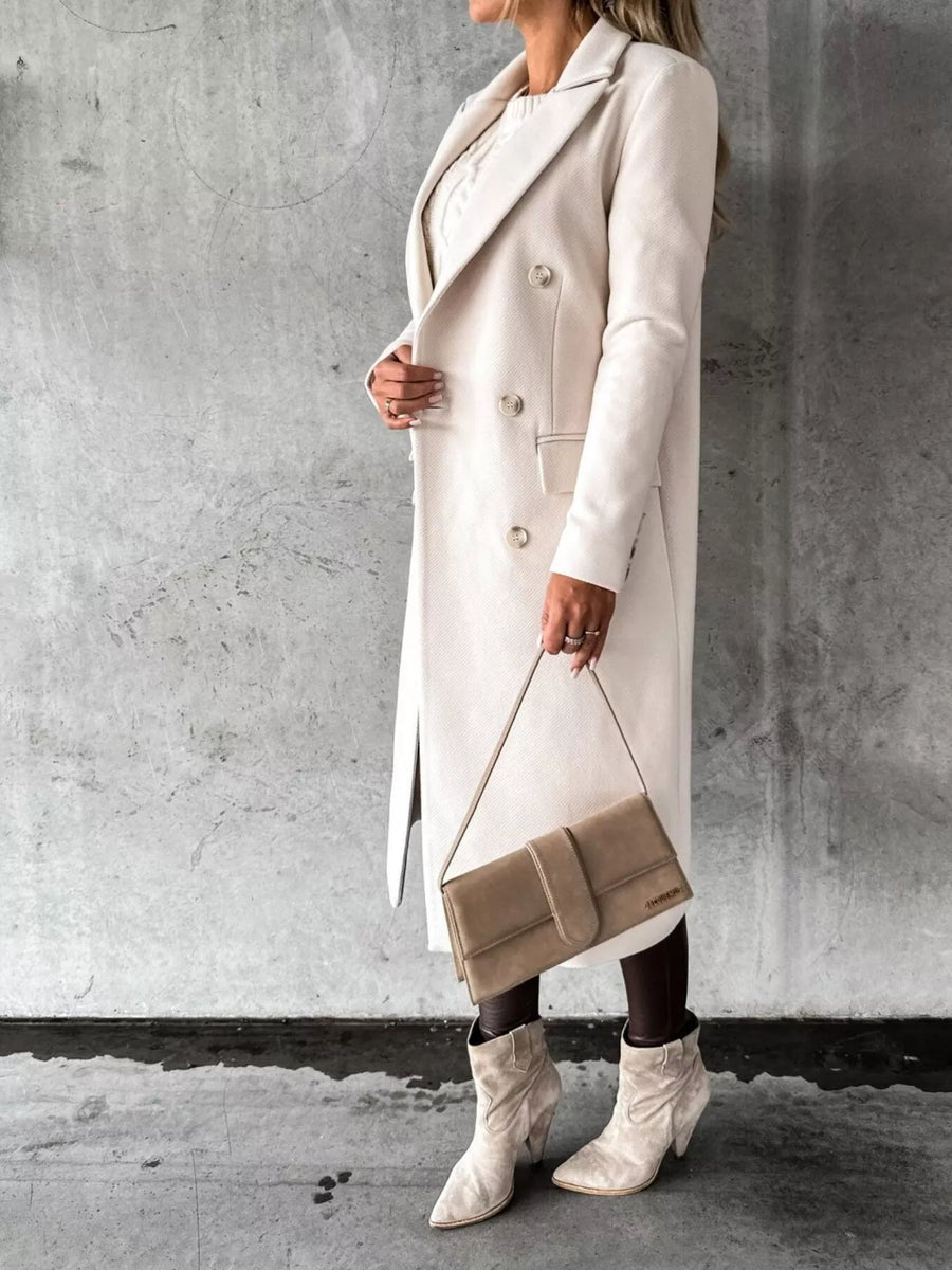Person wearing a long beige coat and white boots, holding a matching handbag against a gray concrete wall.