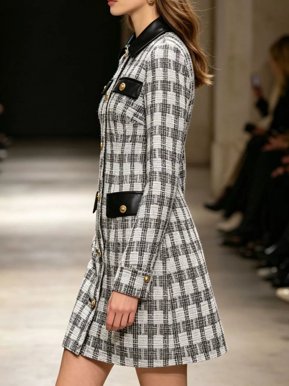 A woman modeling a black and white plaid mini coat with black leather-like trim and gold buttons on a runway.