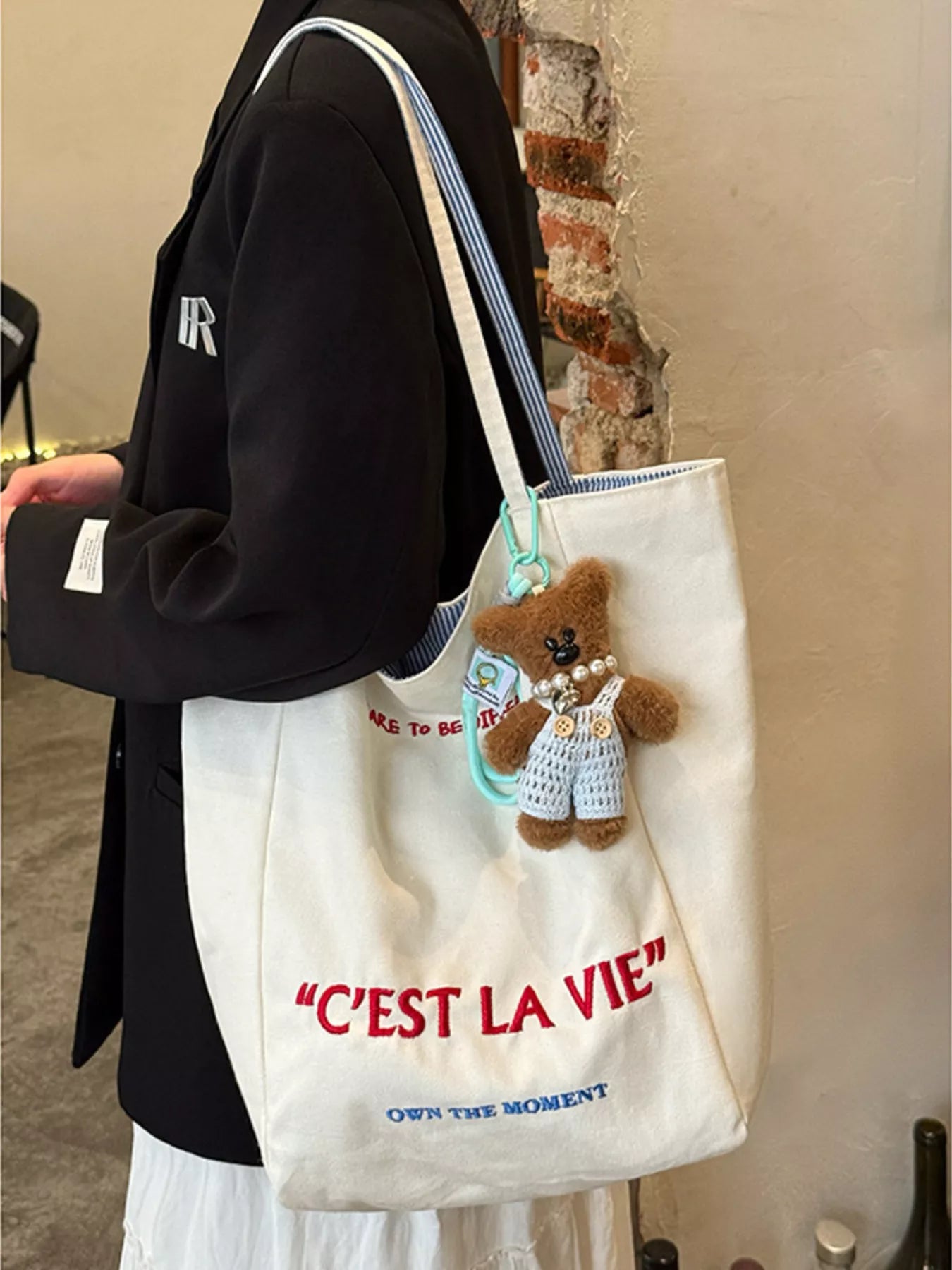 Person holding a white tote bag with a teddy bear charm and 'C'EST LA VIE' text.