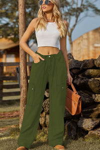 Woman wearing a white top and green pants standing near a stone wall.